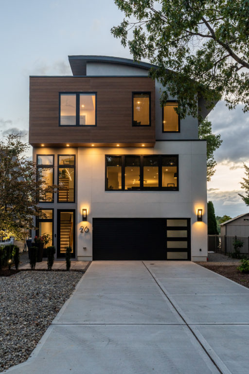 Exterior of a modern home featuring a wide car garage, tall vertical windows, paved driveway, ambient outdoor lighting, and a gravel rock pathway, highlighting contemporary residential design and curb appeal
