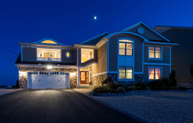 Nighttime exterior of a newly built home with illuminated stone wall, paved driveway, and two barn-door style garages, highlighting modern residential design and curb appeal