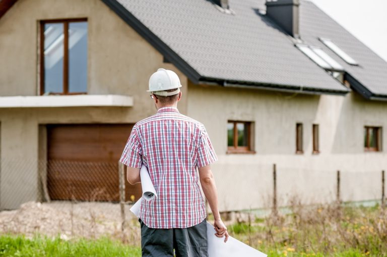 Contractor wearing a hardhat holding design plans walking toward a newly renovated house under construction