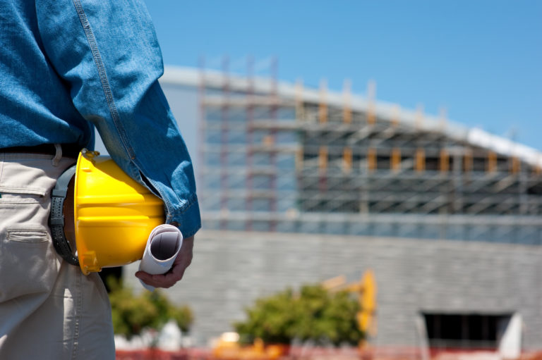 Construction worker holding a yellow hard hat and design plans over looking construction site